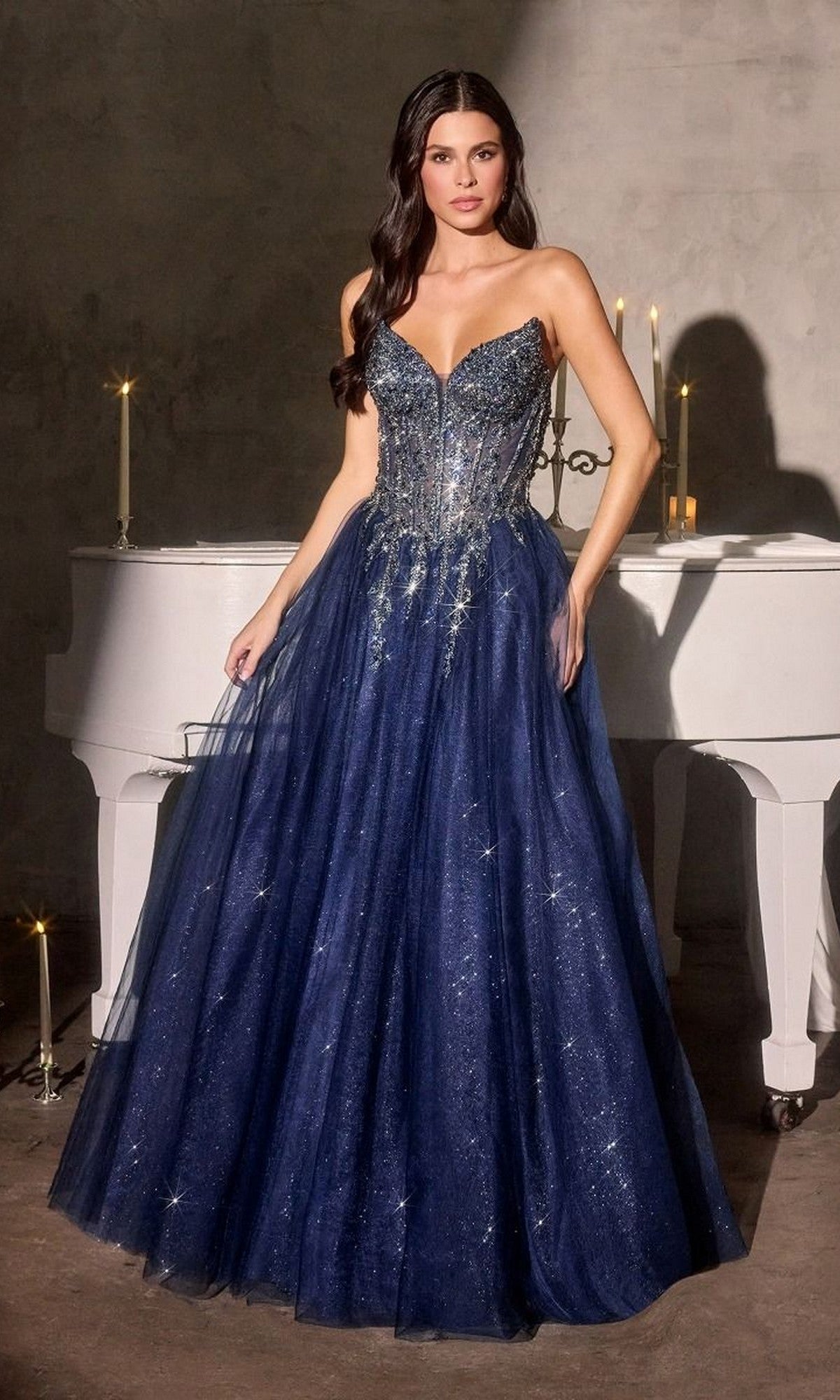 A woman in a Lace-Up Strapless Prom Ball Gown: Ladivine CD721 stands before a white piano with candles, the navy blue glitter tulle dress shimmering elegantly.